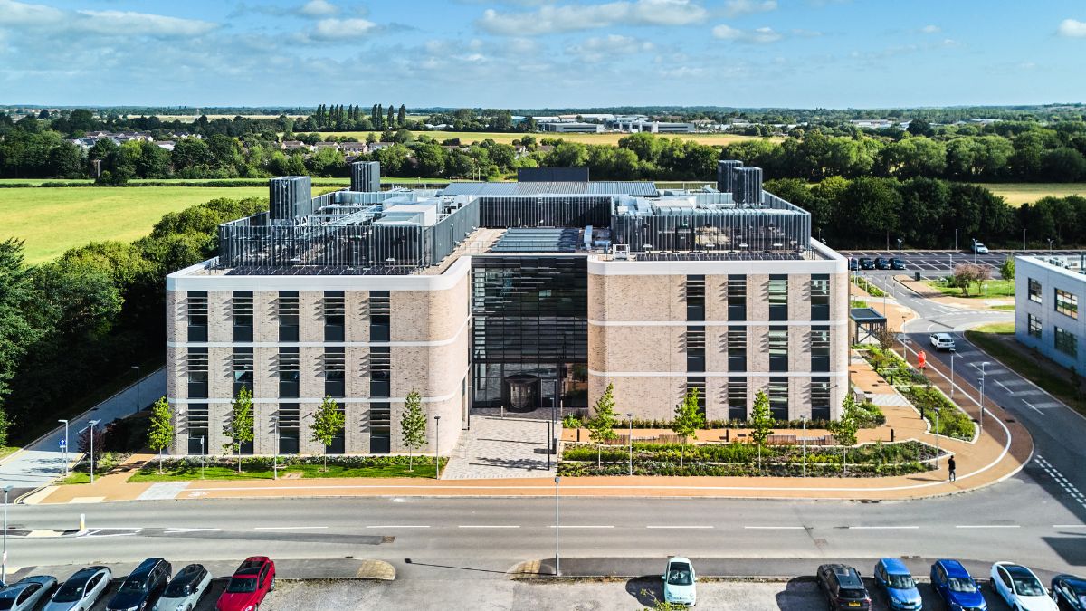 Aerial view of a modern, multi-story commercial building surrounded by greenery and a parking area with parked cars.