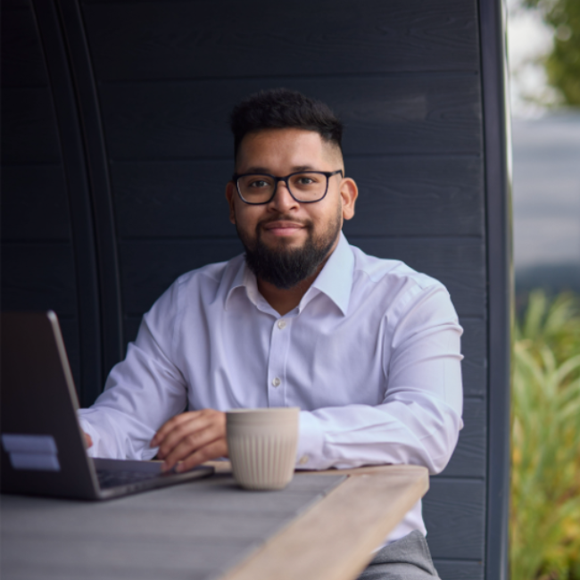 Photo of Karanjit Das sitting with a mug and laptop in front of him whilst he looks at the camera and smiles