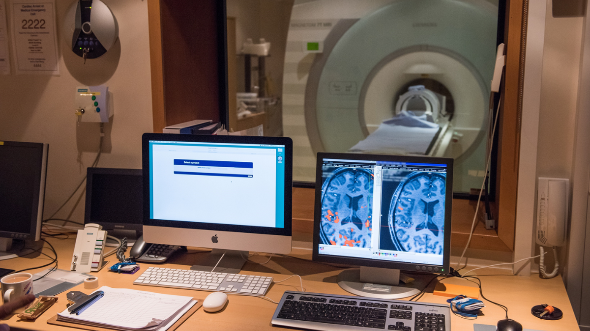 A medical imaging control room featuring two computer monitors displaying brain scan images, a keyboard, and various equipment, with a view of an MRI machine through a large observation window.