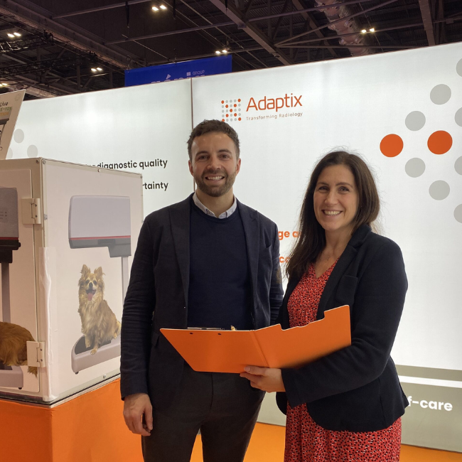 A man and a woman stand in front of a wall with the Adaptix logo. They are both smiling looking at the camera. The woman who is wearing a red dress and black jacket, is holding an orange folder in front of them. The man wears a black jumper and jacket.