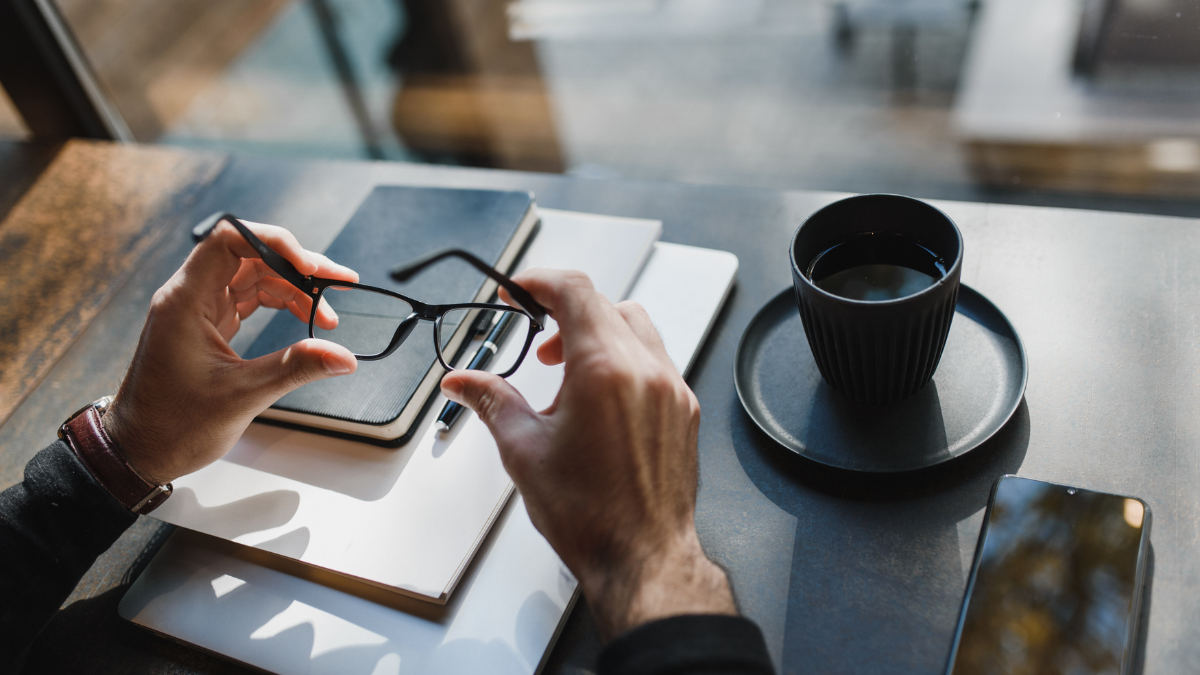 Perspective of a person holding glasses at a desk with a pile of books and coffee cup on the desk.
