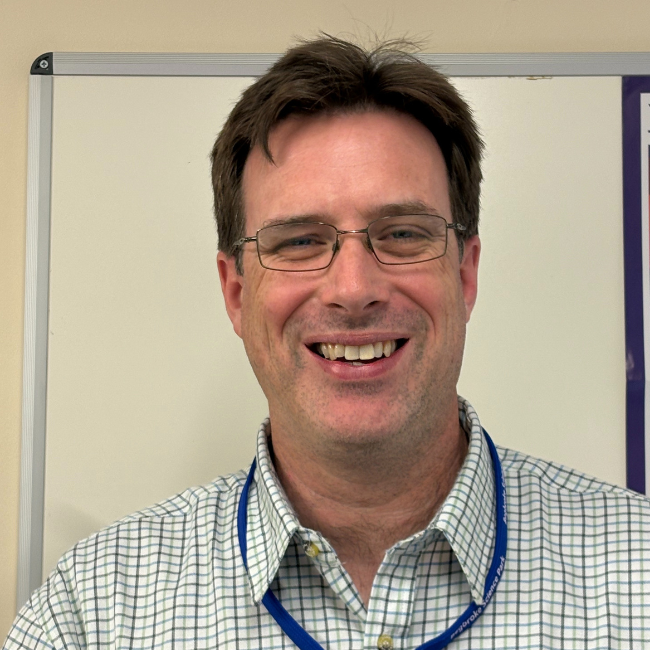 Portrait photo from shoulders up of a man with brown hair standing in front of a white board