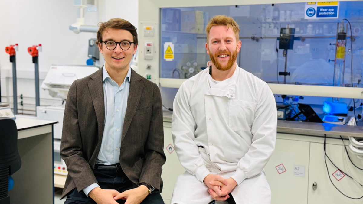 Two men sitting in a lab. One has a suit on and one has a lab coat on.